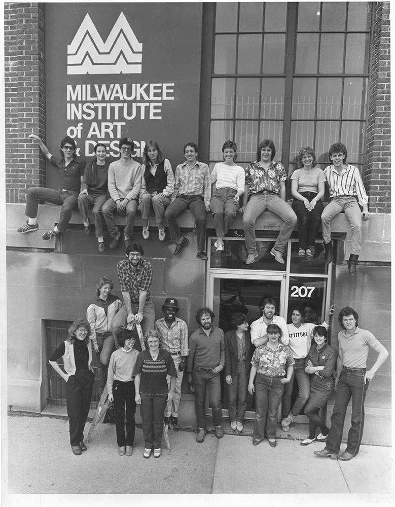 A group of students sits on scaffolding above the door of a MIAD building